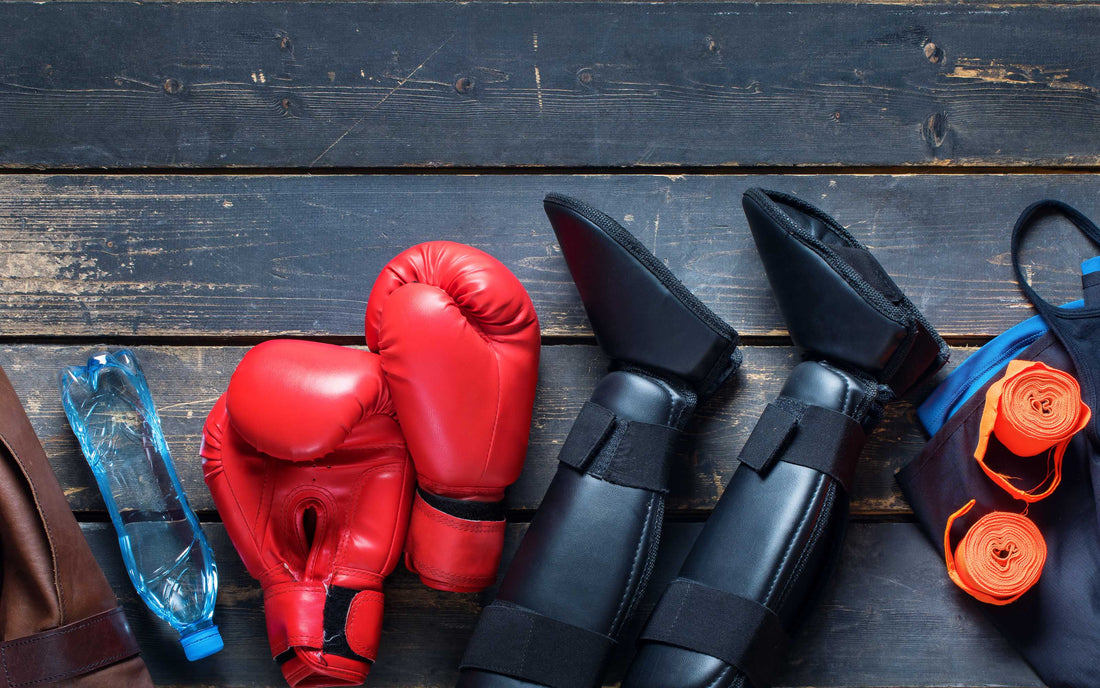Martial arts gear on a wooden surface, including red boxing gloves, black shin guards and foot protectors, orange hand wraps, a blue water bottle, and a blue gym bag.