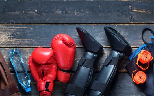 Martial arts gear on a wooden surface, including red boxing gloves, black shin guards and foot protectors, orange hand wraps, a blue water bottle, and a blue gym bag.