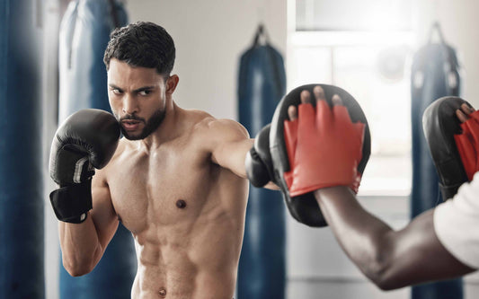 Shirtless man wearing black boxing gloves practices punches with a trainer holding red and black focus mitts in a gym with hanging punching bags in the background