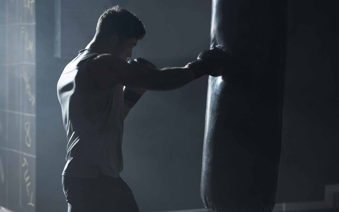 Person wearing boxing gloves and a sleeveless shirt punches a heavy bag in a dimly lit gym, with dramatic lighting highlighting muscle definition and a wall marked with workout notes in the background