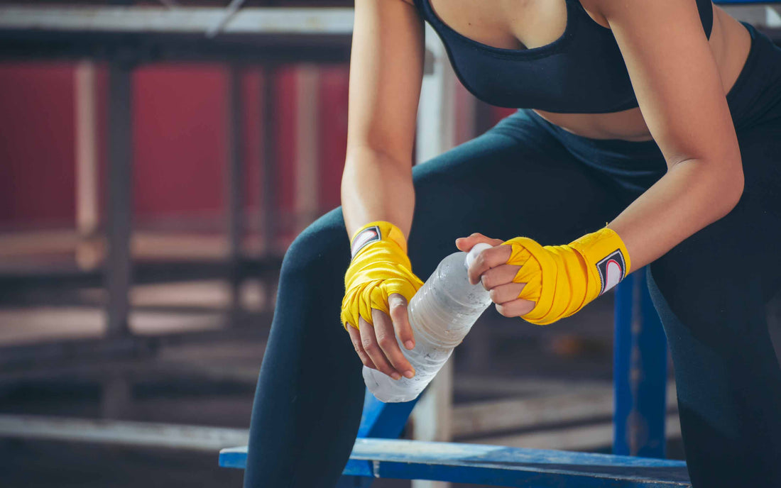 Person in black sportswear with yellow hand wraps holding a water bottle, seated on a blue bench in a gym.