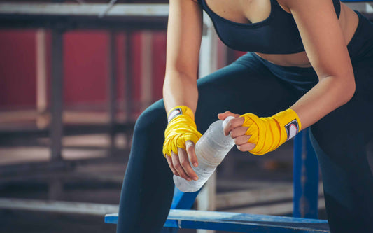 Person in black sportswear with yellow hand wraps holding a water bottle, seated on a blue bench in a gym.