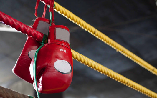 Red boxing gloves hanging by their laces on the ropes of a boxing ring with a dark indoor background