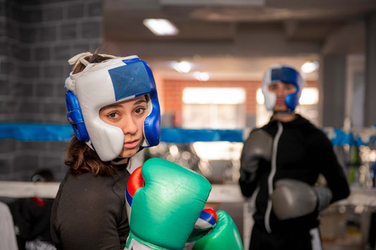 Two people in a boxing gym, wearing protective headgear and gloves, preparing for a sparring session